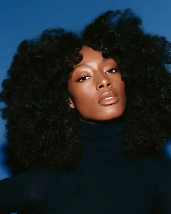 Dense dark afro over a black turtleneck, deep blue backdrop